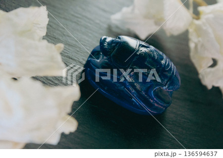Lapis lazuli scarab figurine on dark stone with white petals, ancient Egyptian protection symbol. Lapis lazuli scarab figurine on dark stone with white petals, ancient Egyptian protection symbol. 136594637