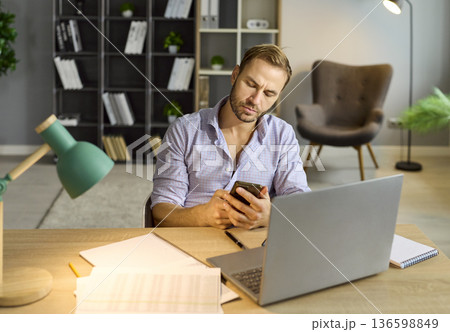 Busy sad man using phone and laptop, suffering from overtime work at office Busy sad man using phone and laptop, suffering from overtime work at office 136598849