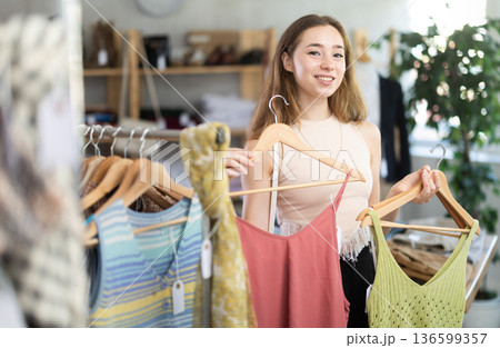 Happy woman chooses a set of clothes for the summer. Visitor to boutique examines new summer collection of clothes 136599357