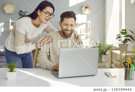 Happy loving woman supporting her husband who is working on his laptop at his desk 136599444