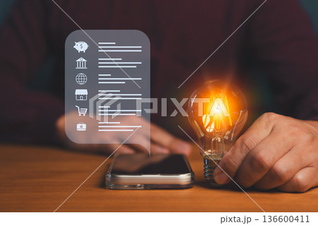 Person holding a glowing light bulb beside a smartphone displaying icons for finance, banking, e-commerce, and business, symbolizing innovation, savings, mobile technology, smart financial solutions. Person holding a glowing light bulb beside a smartphone displaying icons for finance, banking, e-commerce, and business, symbolizing innovation, savings, mobile technology, smart financial solutions. 136600411
