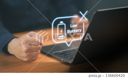 Person using a laptop with low battery warning icon, symbolizing power shortage, technology dependence, computer energy problem, laptop battery issue, energy management, and device usage concept. 136600420