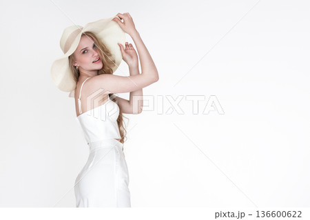 Elegant young blonde woman in chic white summer outfit and straw hat poses against bright, minimalist background. High key lighting creates clean, sophisticated look. Plenty of copy space 136600622