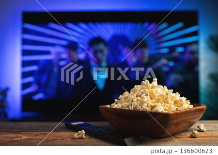 A wooden bowl of popcorn and remote control in the background the TV works. Evening cozy watching a movie or TV series at home A wooden bowl of popcorn and remote control in the background the TV works. Evening cozy watching a movie or TV series at home 136600623