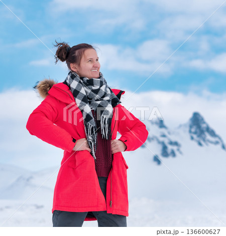 Joyful woman in bright red coat plaid scarf smiles against backdrop of snowy mountains blue sky. Natural daylight lighting. Concept of winter travel, happiness, outdoor adventure 136600627