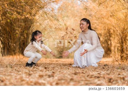 Mother and child girl having fun throwing dry leaves in the air during autumn in golden bamboo forest 136601659