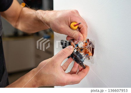 Electrician hands installing electrical light switch with exposed wires into wall socket during home renovation. Concept of electrical work, maintenance, repair and household improvement 136607953