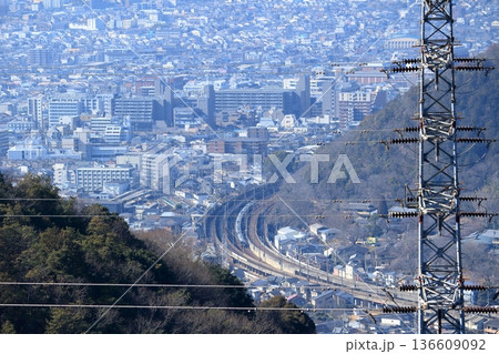 山の中腹に立つ高圧鉄塔越しに見下ろす、山あいを走る列車と街並み 136609092