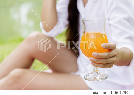 Women hands drink orange juice fresh fruit cool drinking in green park garden. Asian women hands holding healthy drinking glass. Beautiful female pouring orange juice from cocktail bottle outdoors 136612900