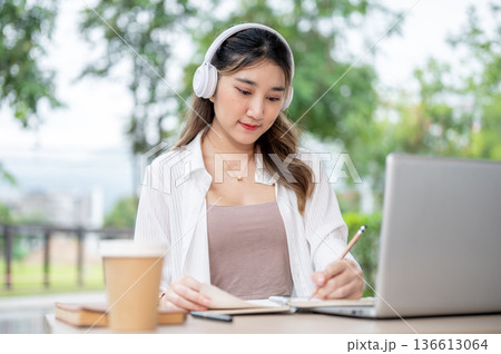 Young asian woman with headphone holding pencil writing in notebook in front of laptop on park table Young asian woman with headphone holding pencil writing in notebook in front of laptop on park table 136613064
