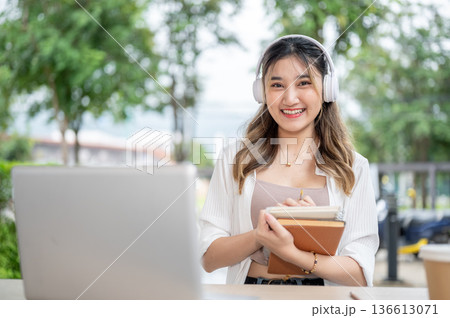 Smiling asian woman with headphones holding pen and books in front of laptop at wooden table in park Smiling asian woman with headphones holding pen and books in front of laptop at wooden table in park 136613071