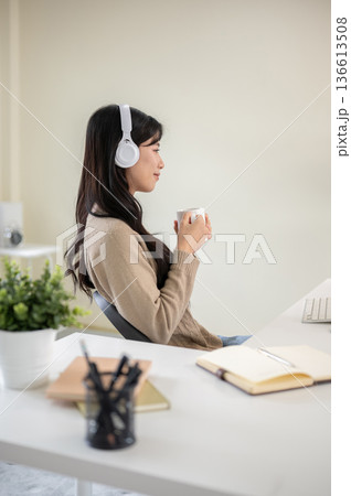 Asian woman wearings headphones holding coffee mug cup while sitting at office table or living room. Asian woman wearings headphones holding coffee mug cup while sitting at office table or living room. 136613508
