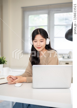 Asian woman wearing headphones looking at laptop on table while sitting in office or living room. 136613521