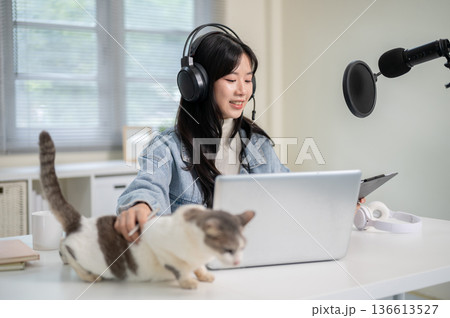 Asian woman patting a cat while hosting a radio or podcast through laptop and mic at home office. 136613527