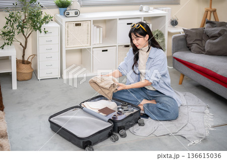 Asian woman putting folded cloth into luggage while organizing or packing in bedroom or living room. 136615036