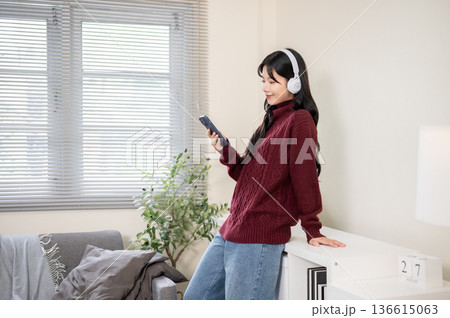 Side view asian woman with headphone holding phone standing leaning against bookshelf in living room 136615063