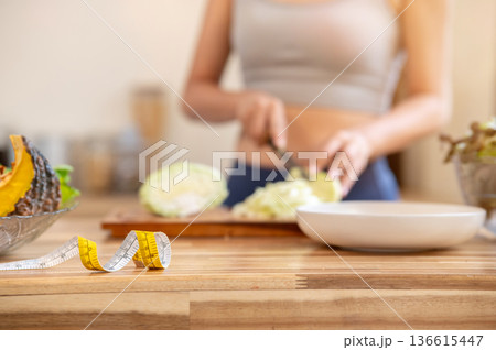 Close up of measuring tape on kitchen cooking counter and blurred sportswear woman cutting vegetable Close up of measuring tape on kitchen cooking counter and blurred sportswear woman cutting vegetable 136615447