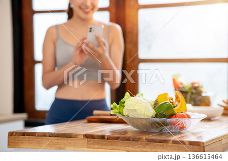 Vegetables in glass bowl on cooking counter and sportswear woman standing holding phone in kitchen. 136615464