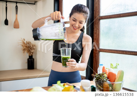 Asian woman in sportswear pouring vegetable juice in blender to a glass standing at kitchen counter. Asian woman in sportswear pouring vegetable juice in blender to a glass standing at kitchen counter. 136615538