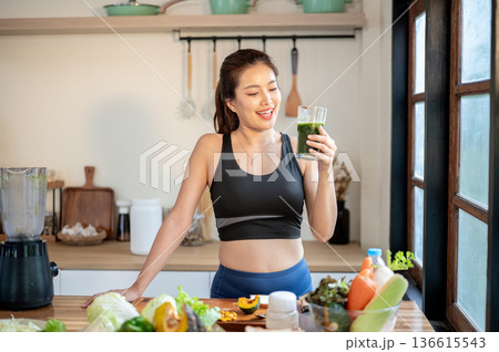Asian sportswear woman looking at vegetable juice glass enjoy drinking standing at kitchen counter. 136615543