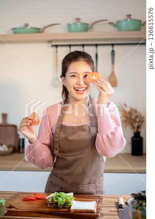 Happy asian woman wearing apron holding tomato slices making a sandwich at kitchen cooking counter. Happy asian woman wearing apron holding tomato slices making a sandwich at kitchen cooking counter. 136615576