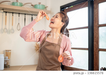 Happy asian woman wearing apron holding and eating tomato slices while cooking at kitchen counter. 136615578