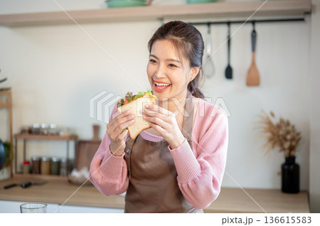 Smiling asian woman wearing apron holding and eating her sandwich making at kitchen cooking counter. 136615583