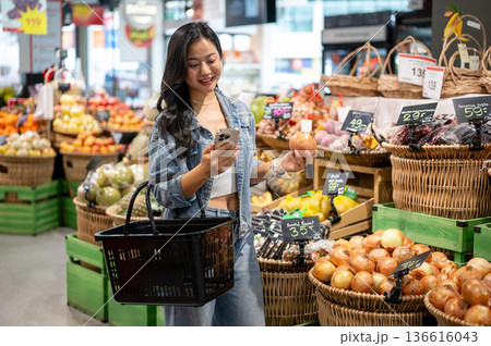 Asian woman holding shopping basket looking at phone while buying onion or vegetables in supermarket Asian woman holding shopping basket looking at phone while buying onion or vegetables in supermarket 136616043