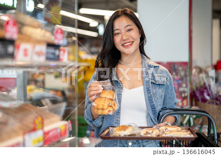 Pretty asian woman holding shopping basket and tray while buying bakery breads in bag at supermarket 136616045