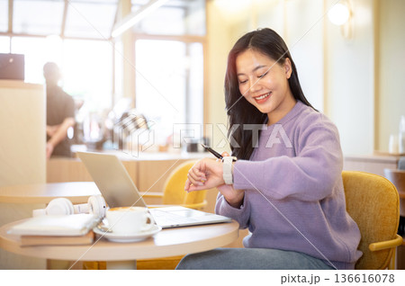 Happy asian woman looking at time on watch and sitting across laptop at table in cafe or coffee shop Happy asian woman looking at time on watch and sitting across laptop at table in cafe or coffee shop 136616078