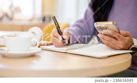 Close up of woman holding pen writing in notebook as looking at phone sitting in cafe or coffee shop Close up of woman holding pen writing in notebook as looking at phone sitting in cafe or coffee shop 136616085