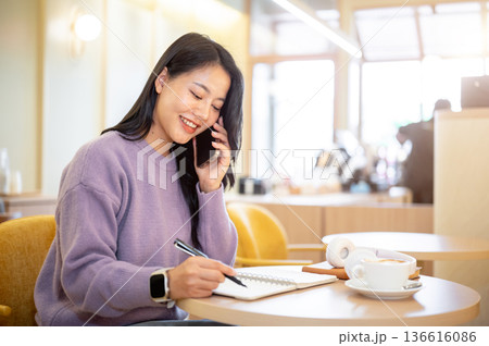 Asian woman holding pen over notebook talking on phone while sitting at table in cafe or coffee shop Asian woman holding pen over notebook talking on phone while sitting at table in cafe or coffee shop 136616086