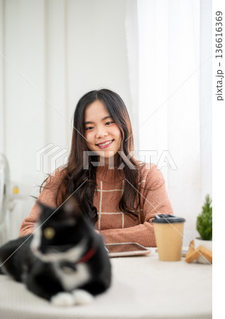 Happy asian woman looking at a black and white fur cat sitting lying on table in living room or cafe 136616369