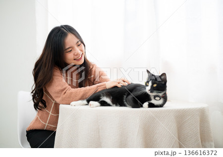 Happy asian woman rubbing fur of a black and white cat sitting lying on table in living room or cafe Happy asian woman rubbing fur of a black and white cat sitting lying on table in living room or cafe 136616372