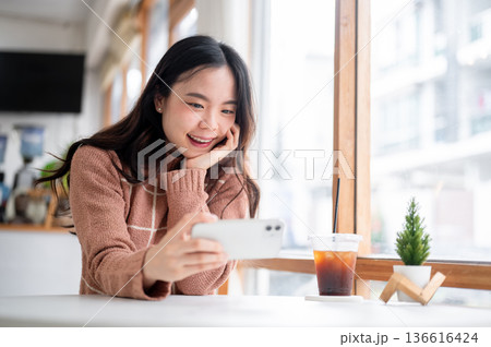 Asian woman holding phone in horizontal watching video at table aside glass wall of cafe coffee shop Asian woman holding phone in horizontal watching video at table aside glass wall of cafe coffee shop 136616424