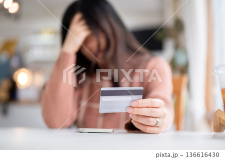 Woman holding head looking at credit card in troubled while sitting at table in cafe or coffee shop. 136616430