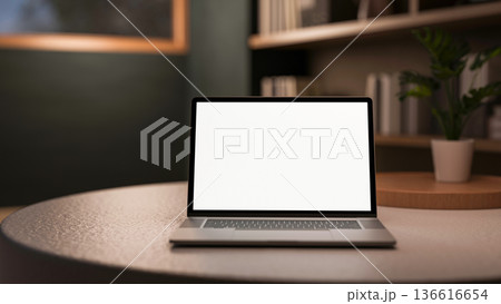 White screen laptop and pot plant on round table aside bookshelf in dim lit living room or library. 136616654