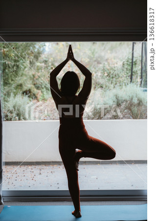 Woman Practicing Yoga in Tree Pose by Window Woman Practicing Yoga in Tree Pose by Window 136618317