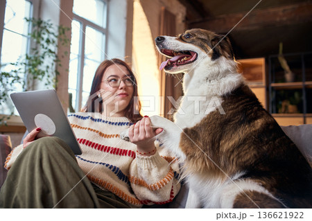 Woman holding dog paw while using tablet on sofa. 136621922