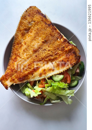 A bowl holds a large piece of fried fish resting on fresh salad with tomatoes, cucumbers, and lettuce. This healthy meal sits on a simple table surface A bowl holds a large piece of fried fish resting on fresh salad with tomatoes, cucumbers, and lettuce. This healthy meal sits on a simple table surface 136621995