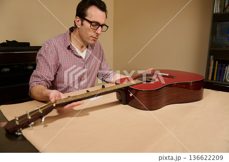 Man adjusting and changing strings on a red acoustic guitar while performing maintenance and repair at a table, demonstrating careful instrument setup and focused craftsmanship. Man adjusting and changing strings on a red acoustic guitar while performing maintenance and repair at a table, demonstrating careful instrument setup and focused craftsmanship. 136622209