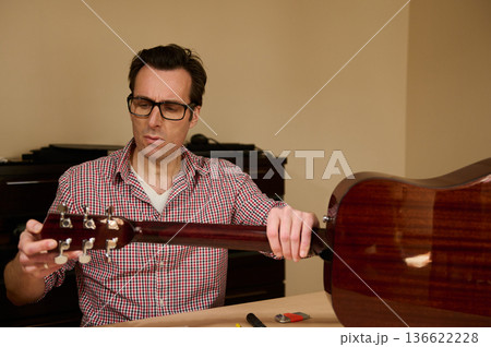 Man at a workbench adjusting and changing guitar strings, demonstrating careful instrument maintenance and repair techniques. Man at a workbench adjusting and changing guitar strings, demonstrating careful instrument maintenance and repair techniques. 136622228