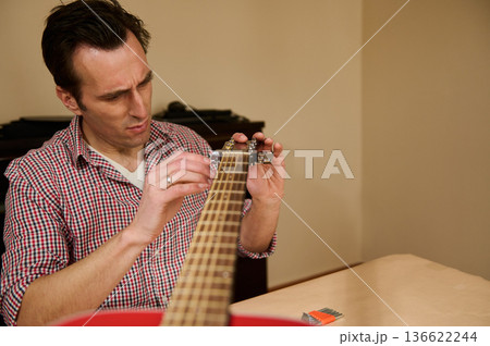 Focused man adjusting and changing guitar strings as part of instrument maintenance and repair, demonstrating tuning, care and hands-on technique in a home or studio setting. Focused man adjusting and changing guitar strings as part of instrument maintenance and repair, demonstrating tuning, care and hands-on technique in a home or studio setting. 136622244