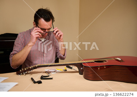 Man adjusting and changing acoustic guitar strings at a table, demonstrating guitar maintenance, repair and tuning in a simple home workshop setting. 136622274