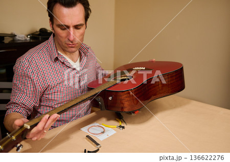 Man inspecting and adjusting acoustic guitar strings on a table, demonstrating careful instrument maintenance and repair with tools for restringing, setup and hobbyist guitar care. Man inspecting and adjusting acoustic guitar strings on a table, demonstrating careful instrument maintenance and repair with tools for restringing, setup and hobbyist guitar care. 136622276