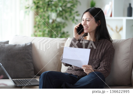Happy asian woman holding letter calling on phone Happy asian woman holding letter calling on phone 136622995
