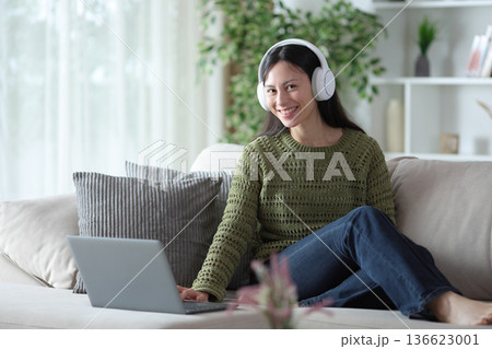 Happy asian woman with headphone and laptop looks at you 136623001