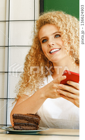 Woman smiles while holding phone near a plate of cake in a cafe Woman smiles while holding phone near a plate of cake in a cafe 136624903