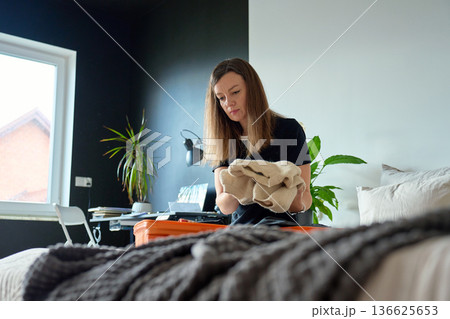 Woman in bedroom holding folded beige sweater over open suitcase Woman in bedroom holding folded beige sweater over open suitcase 136625653