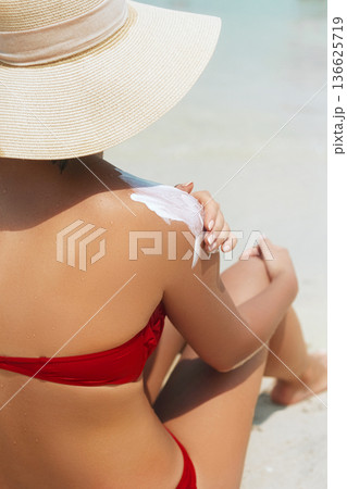 Beautiful woman in hat  sitting on the beach and applying  sunscreen spray on her body.Skincare. Body Sun protection suncream. 136625719